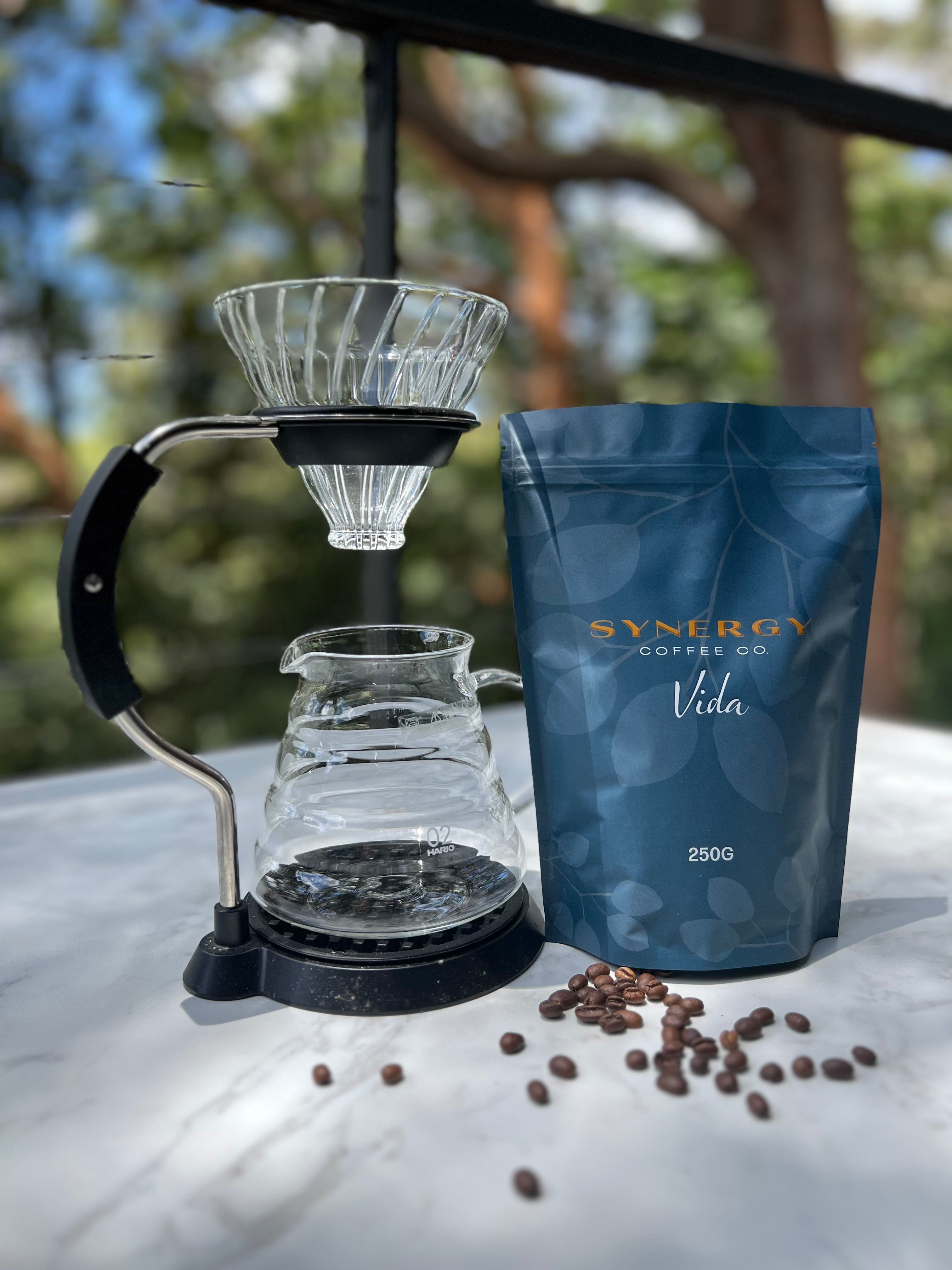 Coffee brewing setup with Synergy Coffee Co. Vida coffee package and scattered coffee beans on a blurred natural background.